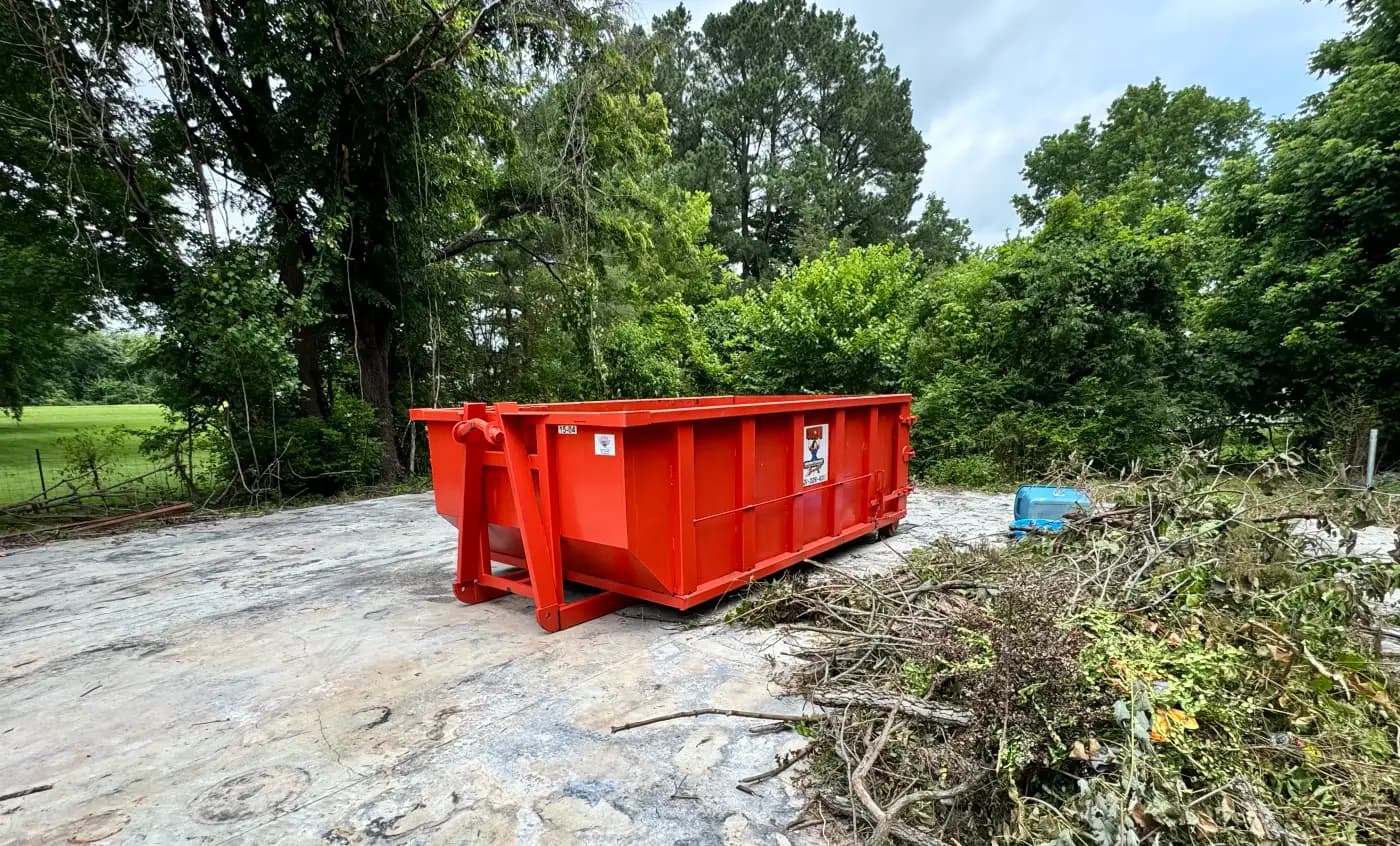 Red Underdog Dumpster 15 yard roll-off container on a concrete pad at a rural property, with a brush pile and an open field and fence line behind it