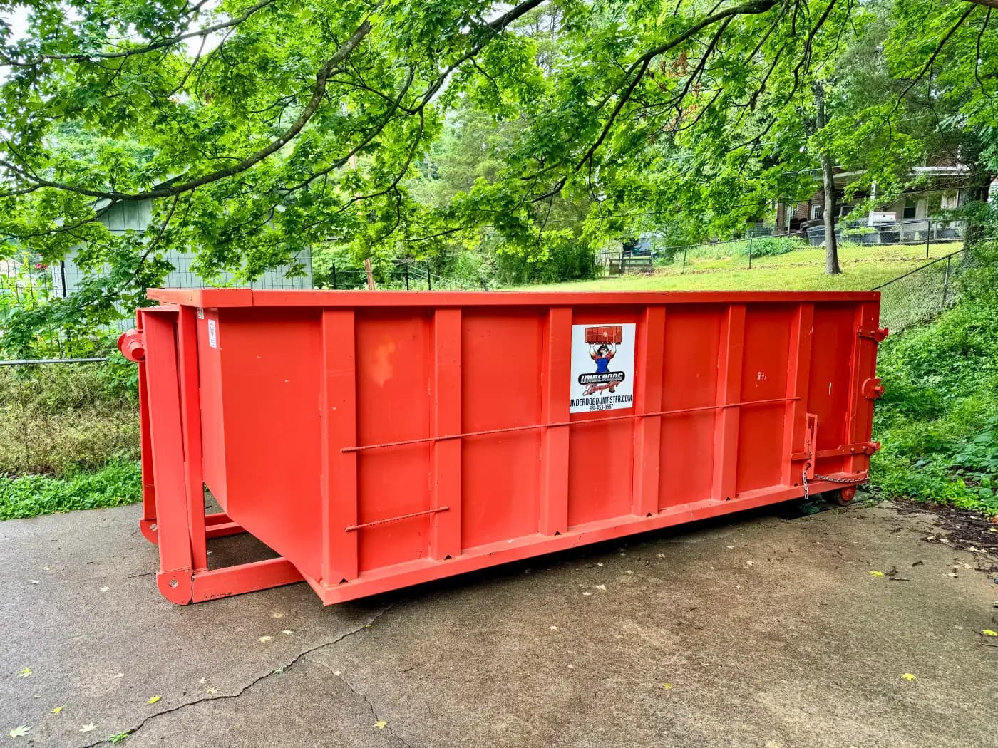 Red Underdog Dumpster 20 yard roll-off container on a residential concrete driveway with tree limbs hanging low overhead