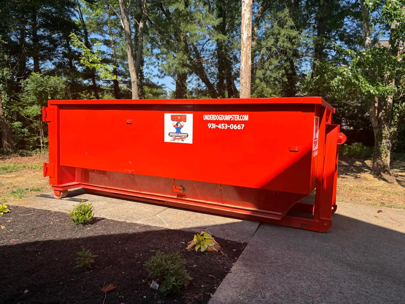 Red Underdog Dumpster 25 yard roll-off container on a concrete driveway next to a mulched landscaping bed, with a protection strip under the rear wheel