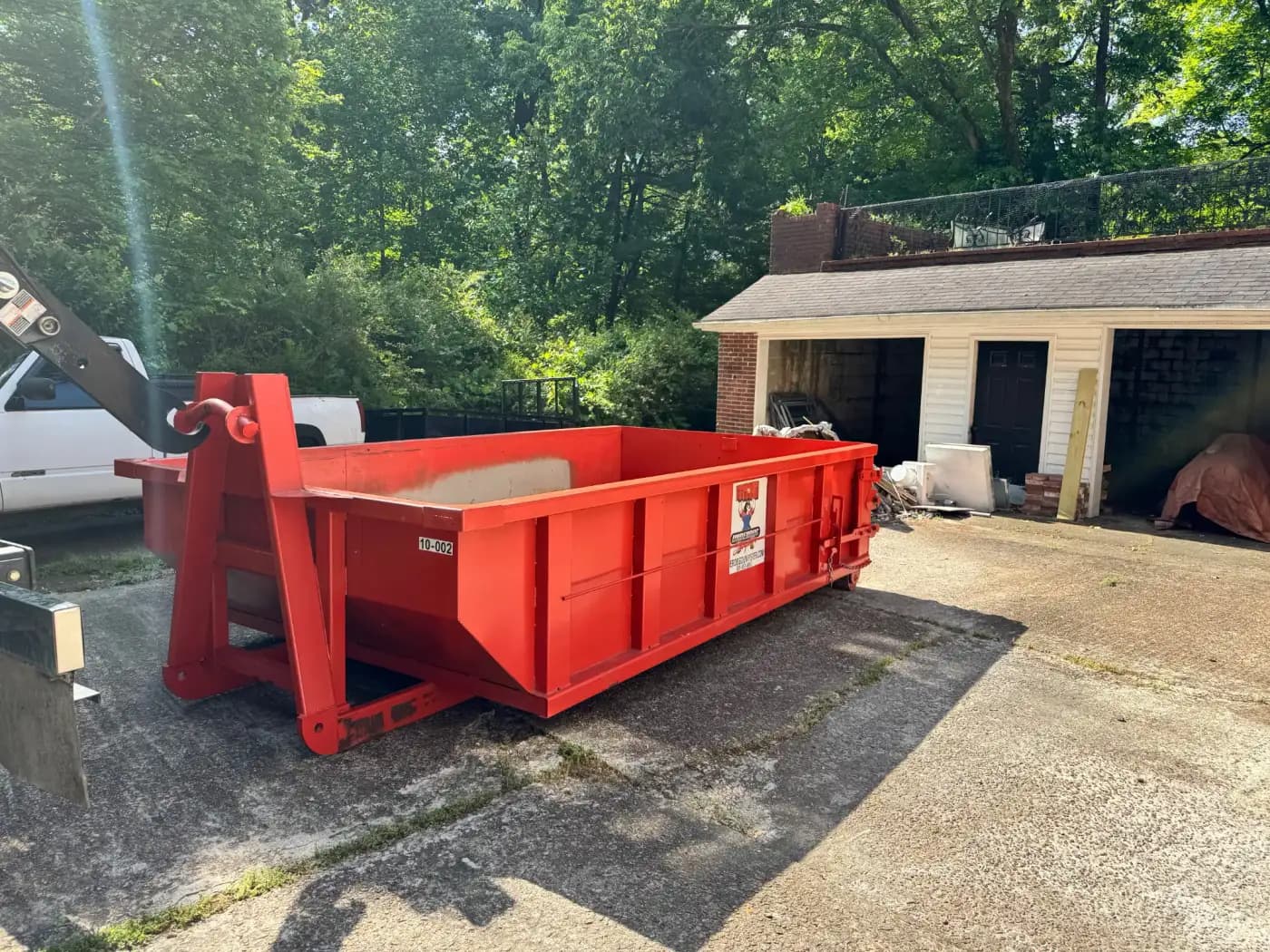 Underdog Dumpster roll-off truck lowering a red 10 yard container onto a concrete driveway beside a detached garage, with the hoist arm still attached